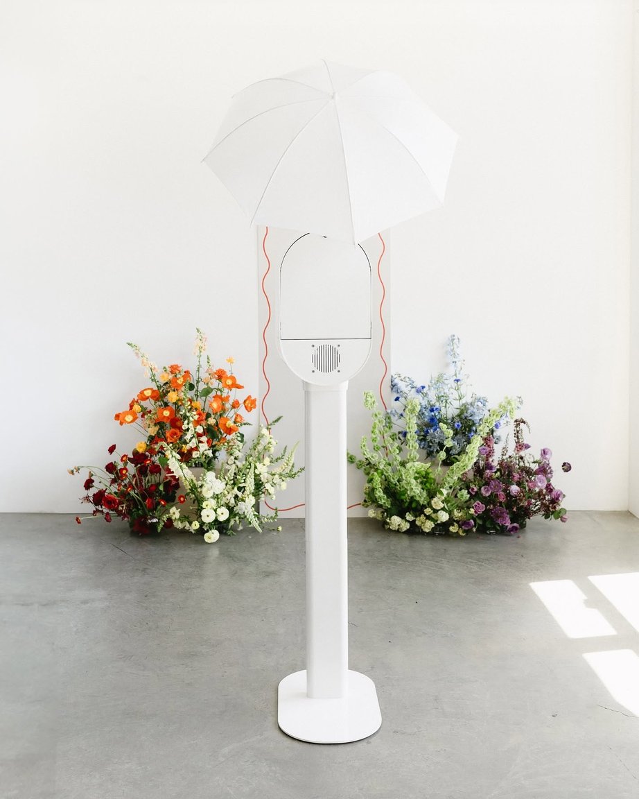 A white photo booth kiosk with umbrella, flowers against white wall