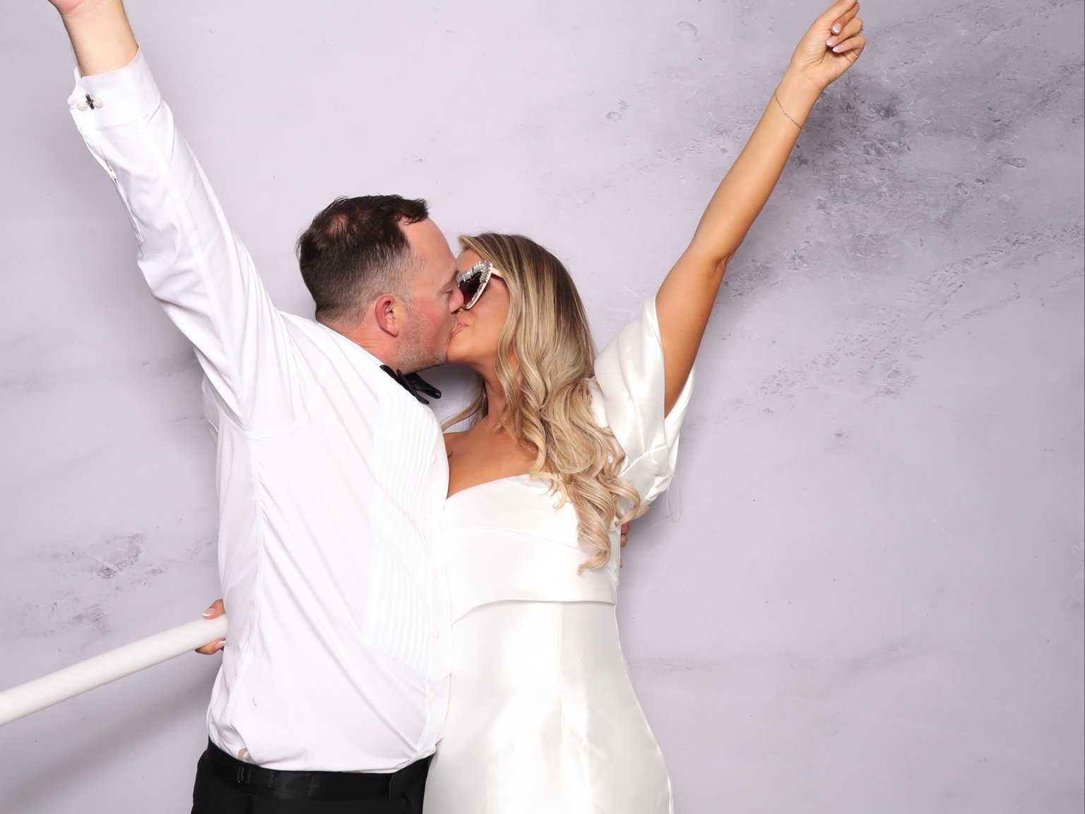 Two people kissing in white outfits, arms raised against light textured backdrop