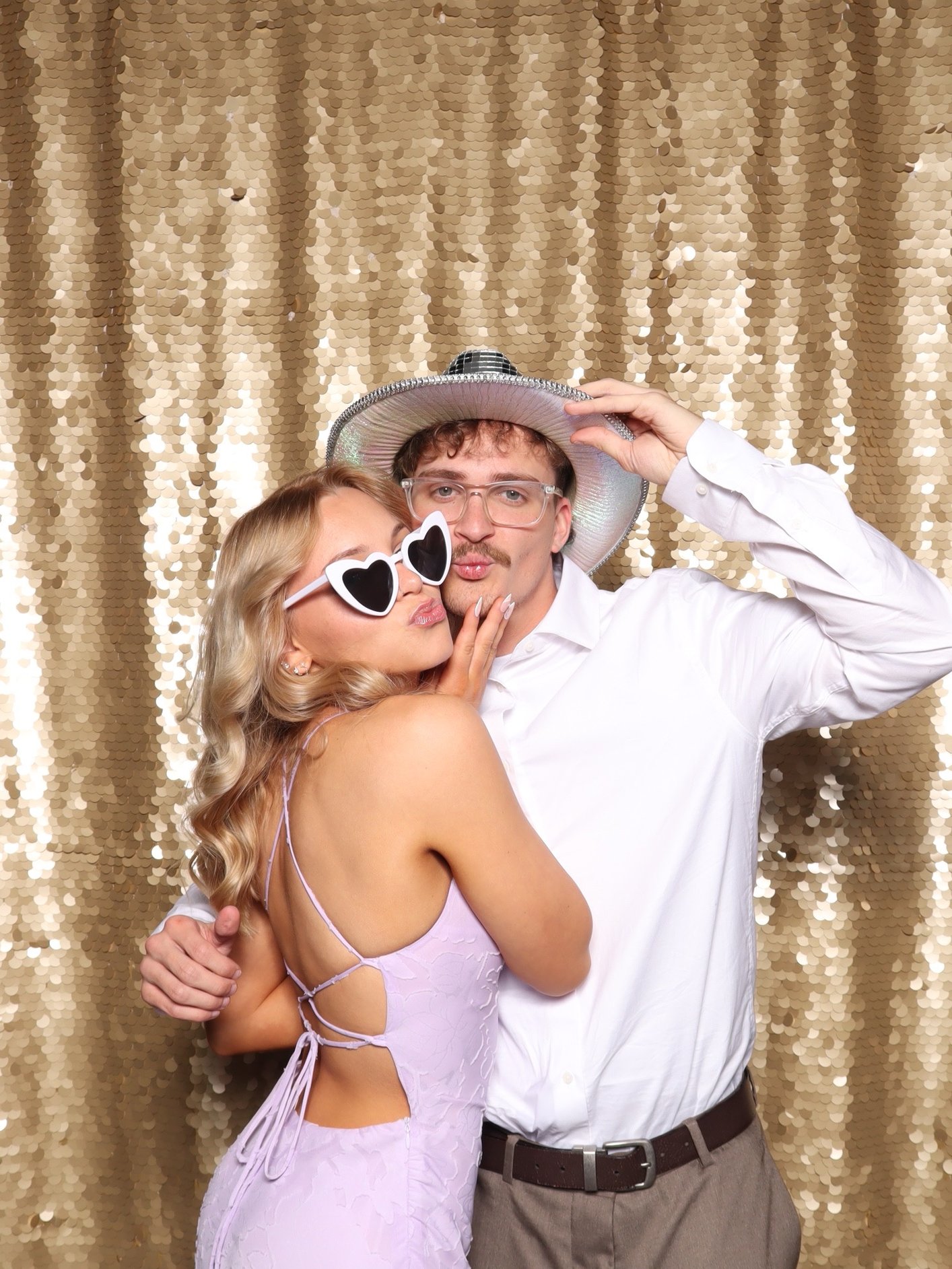 Two people posing in front of a gold sequin backdrop, one wearing heart sunglasses