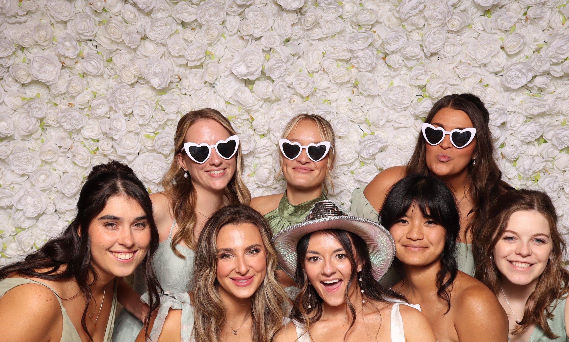 Friends in heart sunglasses and a disco cowboy hat in front of a white flower wall.