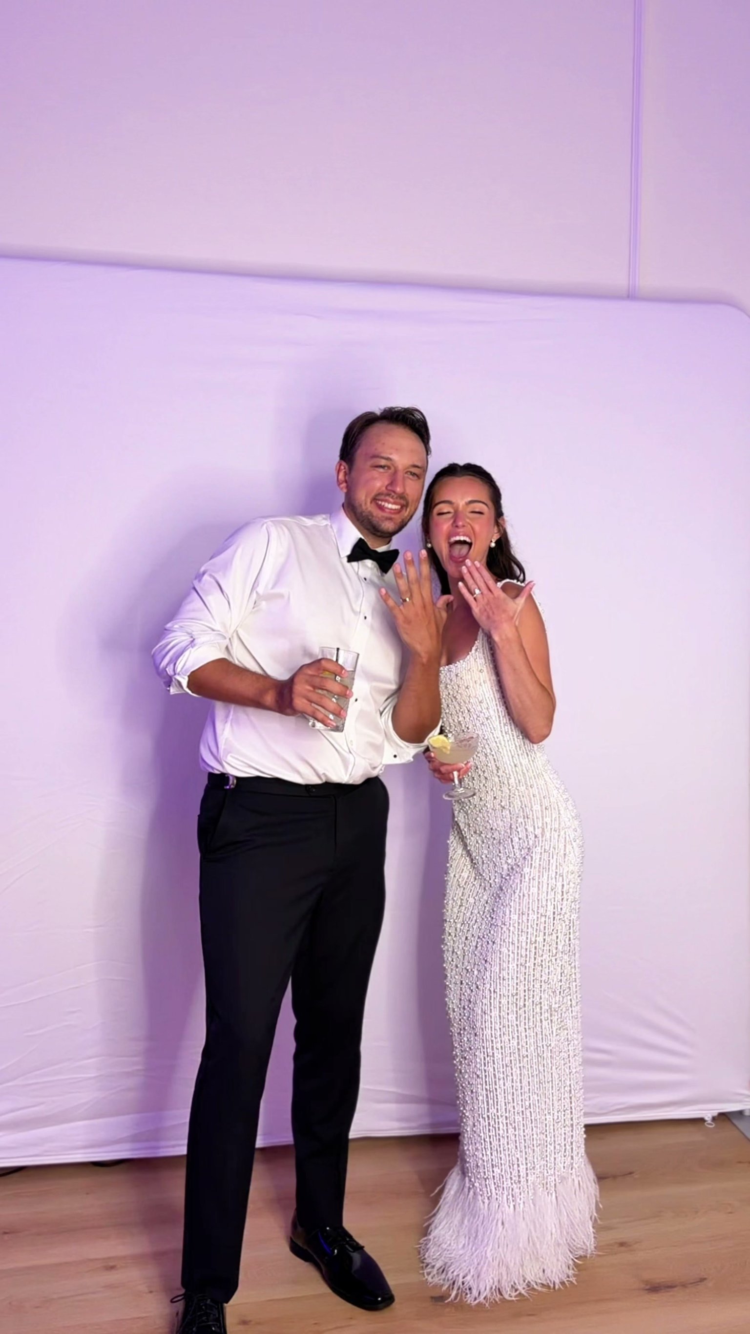 A couple, she in a beaded white gown, at the photo booth backdrop under purple light.